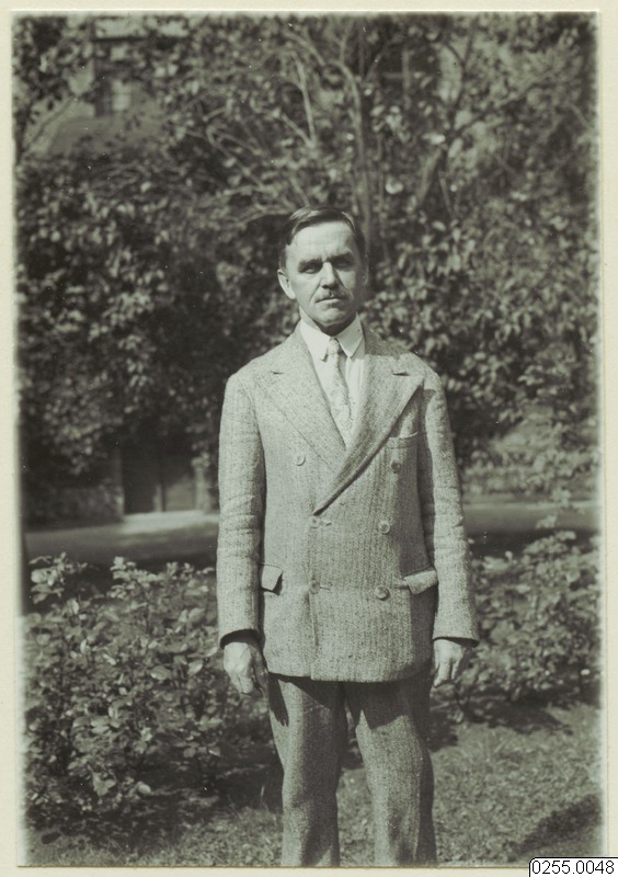 Retrato em preto e branco de Curt Nimuendajú, um homem de meia-idade, em pé, usando um terno de abotoamento duplo e gravata. Ele está posicionado em frente a um jardim com árvores e arbustos.